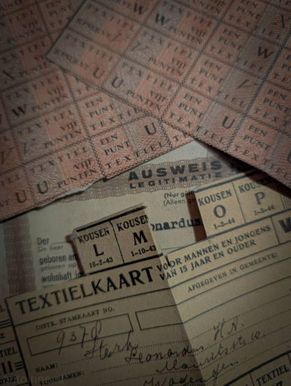 A collection of old documents, primarily related to rationing and identification from World War II during the German occupation of the Netherlands.

The most prominent documents are two reddish-orange sheets and a larger, light brown card in the foreground. These are Textile Ration Coupons. During the occupation, essential goods like clothing and fabrics were scarce and strictly rationed. People needed to present these points to purchase any textile items.

A lighter-colored document is also visible with the German word "AUSWEIS" and the Dutch word "LEGITIMATIE" (Identification).