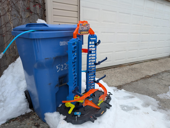 Color photo of a tall plastic Hot Wheels “Ultimate Garage” toy car track standing upright in snow next to a large blue garbage bin, with a white garage door and paved alley in the background.
