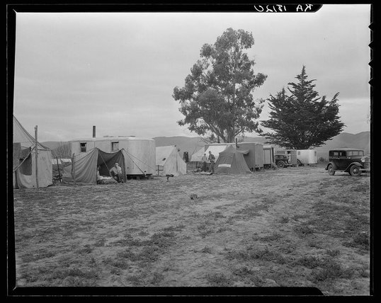 The image depicts a scene with multiple house trailers and tents set up in an outdoor field. The ground appears to be dry grassland, possibly indicative of rural or agricultural surroundings. A few individuals are visible near the tents and one person is inside a tent while another sits on what looks like portable bedding outside their trailer. There's also a vintage vehicle parked nearby with its back facing the camera. Some trees can be seen in the background which suggests this might be located in California as per the provided caption, aligning it to conditions among pea pickers there during that era of time when Dorothea Lange captured such images. The picture is monochromatic and has an old-fashioned quality due to its black-and-white appearance.