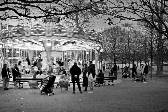 Black and white image of a carousel, on the left, in a public park surrounded by leafless trees.