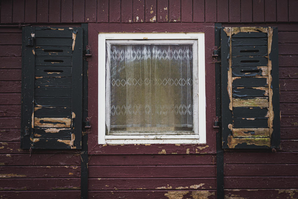 In einer magentaroten Holzwand ist ein weißes, quadartisches Fenster. Links und rechts ist je ein schwarzer Fensterladen angebracht. Im Fenster ist eine Gardine zu sehen. Das Holz ist rissig, die Farbe blättert überall ab. Die Gardine ist grau. Alles vermittelt einen alten und verfallenden Eindruck.