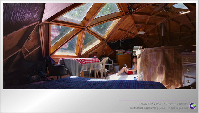 A color indoor photograph. A woman sits on a carpeted floor, typing on a laptop computer. She appears relaxed. The structure she is in has a low, arched ceiling made of striking curved rafters formed into triangles and hexagons. A hexagonal skylight allows sunlight into the room; green pine trees and blue sky are visible through the skylight. There are no animals or vehicles in this picture.