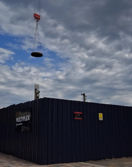 A photo of a construction site with a solid dark metal fence and a crane lifting something overhead. The word Multiplex is visible on the metal fence.