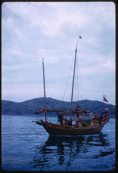 A photograph by Toni Frissel taken as part of a 1959 assignment for Sports Illustrated on sightseeing in Hong Kong. The photo shows an old sailing ship with two tall masts and several people aboard it, some standing at the bow while others are sitting down or leaning against wooden beams along the side. There is one flag flying atop each mast: both have red flags attached to their uppermost sections as they point skyward above dark blue water in Hong Kong harbor. In the background sits a mountain range with various shades of green and brown vegetation, suggesting it's late afternoon based on lighting conditions.

The photograph has been developed from color transparency (slide) format which appears slightly faded or washed out compared to modern-day digital captures but still retains enough detail for interpretation by sighted viewers such as myself. This specific image is part of Frissel’s extensive collection that includes other works, including notable projects in various locations around the world like Hong Kong and New York City where she was based during much of her career spanning over five decades.

For more context about Toni Frissell's life and work please refer to external resources linked at bottom right corner within given source.