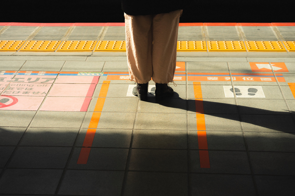 Lower legs of a person in tan pants and black shoes standing on a train platform with orange queue lines, tactile yellow paving near the edge, and a long shadow stretching across tiled ground markings.
