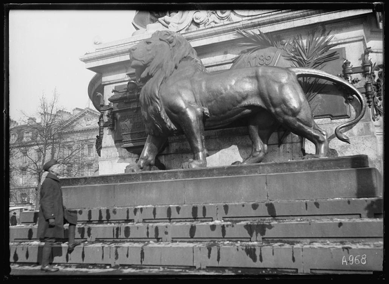 The image depicts a large bronze statue of a lion, prominently displayed on a pedestal with the year 1789 inscribed above it. The lion appears to be roaring and is positioned as if standing guard over its domain. Beside the lion stands an individual in historical clothing, possibly from the late 19th or early 20th century, given their attire which includes a long coat and hat. In the background, there are architectural elements of classical design, including ornate sculptures and detailed masonry work.

The photograph is presented as black-and-white, suggesting it may be an old image capturing historical significance. The setting appears to be outdoors in an urban environment with steps leading up to the pedestal where the lion statue resides. A label "A968" can also be seen on one of the steps, potentially indicating a reference or catalog number for this particular photograph.

Additional context provided mentions that this scene is part of Monument de la Republique by Morice in Paris and refers to it as an image associated with Lewis Hine's work. This suggests historical importance and connection to social reform during his time period.