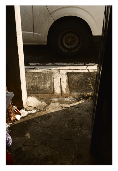 A cramped urban entranceway, dominated by the rear wheel and lower body panel of a dirty white vehicle in the upper section. The foreground features cracked, dark concrete pavement and two rectangular, textured concrete drain covers, dramatically divided by a strong diagonal line of sunlight and shadow. - Google Gemini Latest