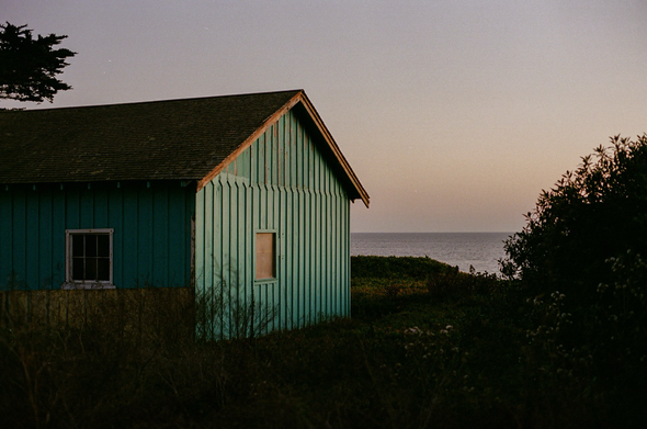 a cabin, painted turquoise with a dark brown slanted roof, faces the ocean over a low cliff at sunset, one side illuminated by the setting sun. trees and bushes appear in silhouette. 