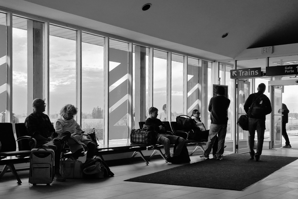 The waiting area at the VIA Rail station. The perspective makes the floor to ceiling windows seem to recede in size from left to right where they meet with a set of doors. The bright early autumn sunshine casts the waiting passengers in shadow, making them almost silhouettes with minimal detail as they wait for the train.