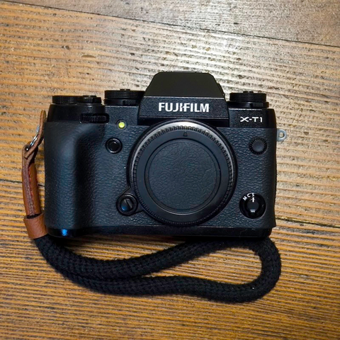 Man sieht eine die Frontseite einer Fujifilm X-T1 die auf einem Holzboden liegt Diese hat eine schwarze Handschlaufe.