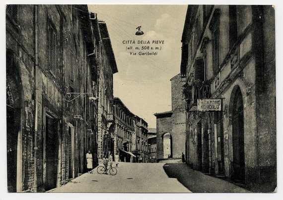 The image depicts a historic street scene from Città della Pieve, Italy. This monochrome photograph shows an alleyway with buildings on either side that exhibit traditional architectural features such as shuttered windows and ornate balconies. The height of the structures is marked at approximately 508 meters above sea level.

In the foreground, a person is walking while another individual rides a bicycle along the street. Various signs are attached to the walls; one reads "Vicino al Pieve," suggesting proximity to a significant religious structure or church (pive typically refers to churches in Italian contexts). A hanging sign with text that translates as 'Nearby Church' indicates this area's association with religion.

The image has an atmospheric quality, likely due to its age and the use of black-and-white film. The caption also mentions "Città della Pieve" prominently at the top, which is identified in a foreign language below it. A reference is made to Garibaldi Street via, possibly indicating this street's connection or relevance to Giuseppe Garibaldi, an Italian general and politician.

Overall, the image conveys a sense of historical charm and everyday life within a European town during what appears to be early 20th-century times.