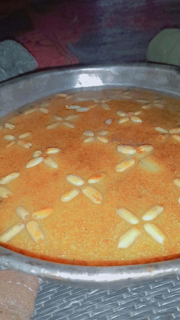 A close-up shot of a round, golden-brown dessert. It's a semolina cake (Basbousa), decorated with symmetrical arrangements of toasted almond slices in a pattern resembling flowers, resting in a silver, shallow pan.