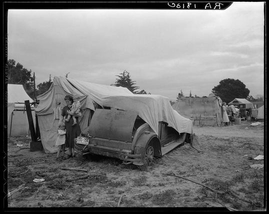 The image is a black and white photograph depicting an outdoor scene with several elements. At the center, there's a vintage car partially covered by what appears to be a tarp or sheet. The vehicle shows signs of wear and rust, suggesting it has been exposed to harsh conditions for some time.

Beside this car stands a woman wearing long-sleeve garments which seem worn out and practical. She is holding onto the door frame with one hand while her other arm supports what appears to be two children; possibly infants or toddlers dressed in simple clothing appropriate for outdoor work. The environment suggests an area of temporary settlement, perhaps related to agricultural labor.

In the background, there are several tents set up on a rough terrain which indicates that this might be a campsite of migrant workers. Some trees can be seen further back, and various items like buckets or containers are scattered around, hinting at daily life activities in such settlements. The sky is overcast with no visible sunlight.

The photograph carries an air of hardship but also resilience as indicated by the presence of this family amidst their surroundings. It's a poignant snapshot capturing themes often associated with migration—struggle and endurance against adversity for sustenance.