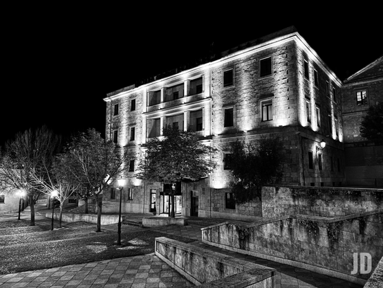 Una imagen en blanco y negro de un gran edificio de piedra por la noche, destacando su arquitectura. El edificio de tres o cuatro pisos está fuertemente iluminado desde abajo, lo que resalta la textura de sus muros y sus múltiples ventanas oscuras, así como los balcones en los pisos superiores. En primer plano y a la izquierda, hay varios árboles con follaje escaso o ramas desnudas, iluminados por farolas que proyectan círculos de luz en el suelo adoquinado y las zonas ajardinadas. A la derecha, se aprecian muros bajos y escaleras de piedra que descienden, creando diferentes niveles en el entorno. El cielo es completamente oscuro.