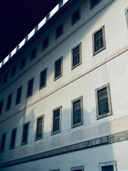 Fachada con ventanas rectangulares cubiertas de verjas, parcialmente en sombras. Es el Museo Reina Sofía en Madrid.