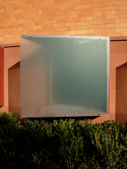 Color photo of a window which extends out at an angle from the wall plane. To enhance privacy, the single square pane of glass has been etched with a series of wide, horizontal stripes. The masonry wall in which the window is set is composed of blocks of pink stone (lower section) and orange bricks (upper section). There is shrubbery beneath the window.