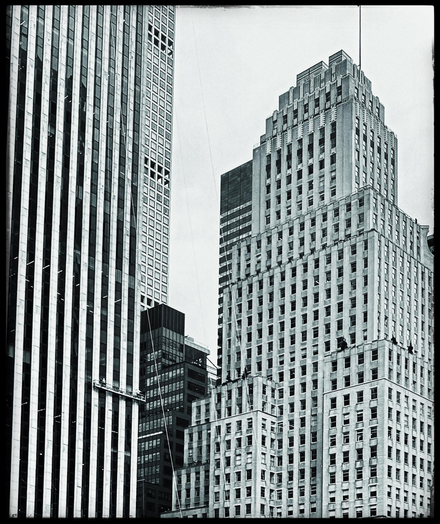 A black-and-white photo taken primarily of two buildings that could be seen from 59th St. facing west. The building on the left is Modern, very tall and the architecture of the windows almost makes it look like vertical stripes of light and dark. On the bottom third of the photo, on the building, can be seen a long platform hanging down with people on it cleaning the windows. The building on the right is an older style building that is white with so many small windows it would be impossible to count. The building has several layers to it getting slightly smaller as it goes up. On one of the parapets are some black spots that were also people either doing work on the building or cleaning windows. There is a smaller black building in between them and the sky is a plain gray. The photo has a black frame