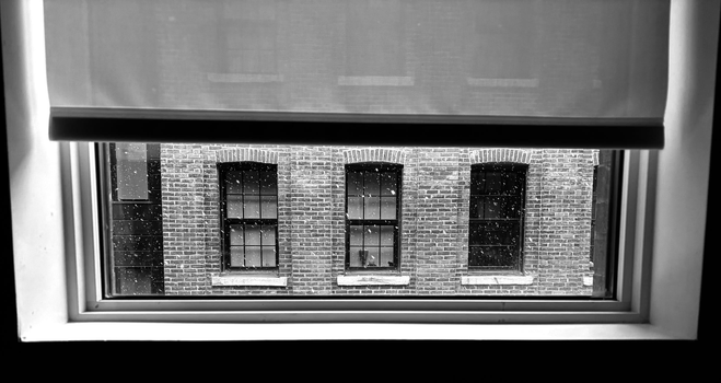A black and white photograph of three side-by-side windows, framed in black, in a brick wall, with falling snow. The photo was taken from the inside of the window in the building opposite, with the shade lowered almost to the bottom, creating a soft, diffused light within a simple white frame. Three other similar windows in the opposite wall on the exterior are visible through the shade, above the first three.

Photographie noir et blanc de trois fenêtres côte à côte, au cadrage noir, dans un mur de briques, avec la neige qui tombe. La photo est prise par la fenêtre à l'intérieur du bâtiment en face, avec la toile baissée presque jusqu'en bas, créant une lumière tamisée, à l'intérieur d'un cadre blanc et simple. Trois autres fenêtres semblables dans le mur d'en face à l'extérieur sont visibles  à travers la toile, en haut des trois premières.
