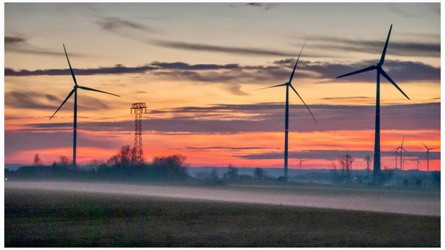 A serene landscape featuring three wind turbines silhouetted against a colorful sunset. A power line tower is visible, with mist rising from a field in the foreground.