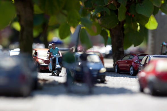 A miniature scene featuring a person on a scooter navigating through a street full of miniature cars with trees overhead. The focus is on the central figure, with a shallow depth of field blurring the foreground and background.