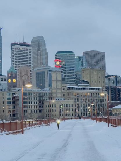 As dusk begins lights along a bridge are illuminated. The snow covered bridge top has only people walking and running. One person running has a light. A city skyline stands beyond. One prominent sign on a building says ‘Northstar Blankets’