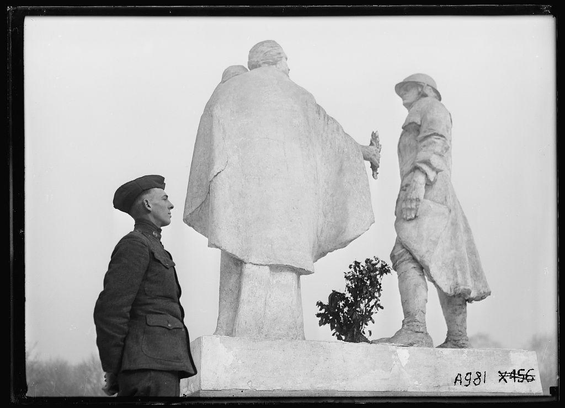 The image depicts a statue featuring three figures; one larger figure with an outstretched arm, seemingly offering something to the other two smaller statues. The larger statue is draped in what appears to be a cloak or coat and has a bald head, while the other two are dressed more formally.

In front of this statue stands a man wearing a military uniform from the early 20th century, possibly World War I era based on his attire which includes a peaked cap. His posture suggests he is looking at the statue with interest or reflection.

The image appears to be in black and white, indicating it may have been taken during that time period or has been edited to give an older appearance. There's also some handwritten text near the bottom right corner of the photo: "A981 X456."

Given the military uniform and historical context suggested by this photograph, it is likely commemorating soldiers from World War I in France. The statue may represent a tribute or memorial for fallen troops during that war.

The image also has some text at its bottom which reads: 'Paris.' This could imply that the photo was taken near Paris, possibly where such statues are often found along with other historical monuments and landmarks of significance to military history from WWI.

Additional information about this specific statue can be researched further. The mention in Paris indicates a possible conn [...]