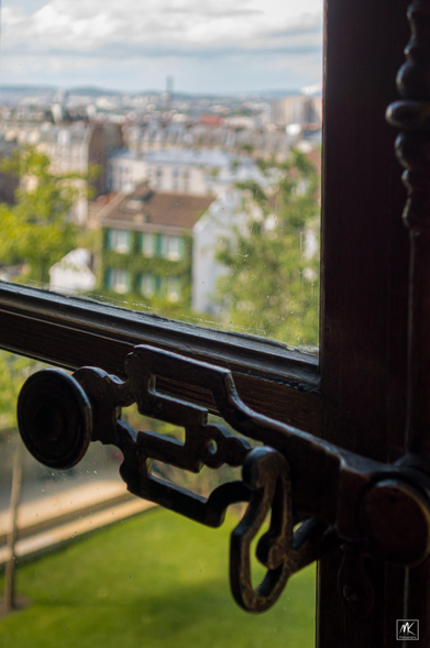 Close up color photo of an antique window latch mechanism and a view out the window behind it of the buildings of Montmartre. 