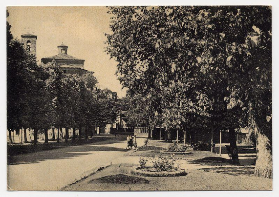 The image depicts a black and white scene of an outdoor park or garden area. In the foreground, there is a paved path with some bushes on either side. On this path, two individuals appear to be walking away from the viewer's perspective.

To the left in the background stands a building featuring traditional architectural elements such as arched windows and what appears to be a tower structure with bell openings at different levels. Adjacent to it is another large building with a domed roof, possibly indicating an institutional or historical significance.

The middle ground shows more trees that form a dense canopy over much of the area, providing shade on the path below. The overall atmosphere suggests tranquility and perhaps a leisurely stroll through this serene urban green space.