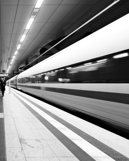Auf dem Schwarzweißfoto fährt ein Hochgeschwindigkeitszug in einer Bahnhofshalle an einem Bahnsteig entlang. Der Betrachter steht auf dem Bahnsteig, der Blick folgt dem Bahnsteig. Die Deckenleuchten, der Zug, das Fensterband und ein Längsstreifen auf dem Zug, die Bahnsteigkante und ein Leitstreifen – alle Linien führen auf einen Punkt mittig am linken Bildrand. Dort läuft eine Person den Bahnsteig entlang, dem Betrachter entgegen.

In the black-and-white photo, a high-speed train travels along a platform in a station concourse. The viewer is standing on the platform, their gaze following the platform. The ceiling lights, the train, the row of windows and a longitudinal stripe on the train, the edge of the platform and a guide strip – all lines lead to a point in the center of the left edge of the image. There, a person is walking along the platform, towards the viewer.