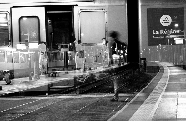 Une photo en noir et blanc au format paysage. Exposition multiple
Un quai de gare avec ses lampadaires et un train à l'arrêt, s'étire de la gauche vers la droite, se perd dans la nuit et constitue le fond de l'image  Une seconde prise montre un wagon, porte ouverte et disposé perpendiculairement au quai de l'image de fond. Au premier plan, et un peu sur la droite du centre, un personnage traversé de lumière semble hésiter à monter dans le train.