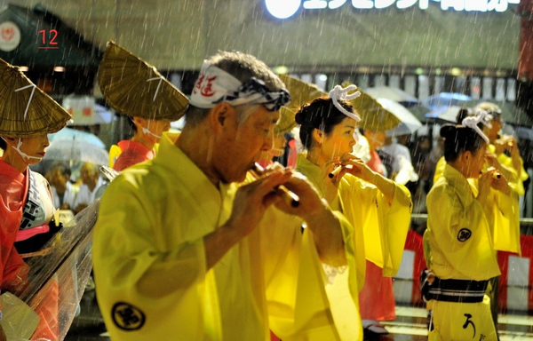 Photo couleur d'un festival traditionnel au japon, sous la grosse pluie. Des participants défilent en kimono dans la rue. Des participants sont en kimono jaune et jouent de la flûte suivient par des femmes portant un kimono orange-rouge et un chapeau traditionnel en paille. Elles jouent du shamisen, instrument à cordes japonais.