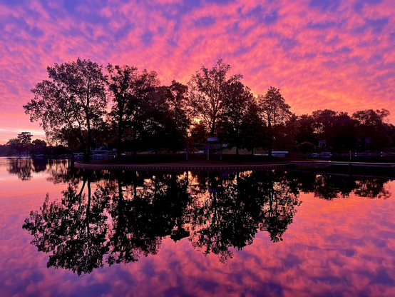 Photo of a colorful red, orange, pink, and purple sunrise over a lake with the clouds reflected in the lake and an island with trees in silhouette in the center.