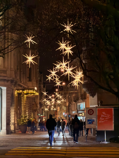 Eine geschäftige Stadtstraße bei Nacht, geschmückt mit hellen sternförmigen Lichtern. Die Menschen gehen den Bürgersteig entlang, und festliche Dekorationen sind auf den Gebäuden sichtbar, was eine fröhliche Atmosphäre schafft. Straßenschilder und Schaufenster sind beleuchtet und tragen zur pulsierenden Szene bei.