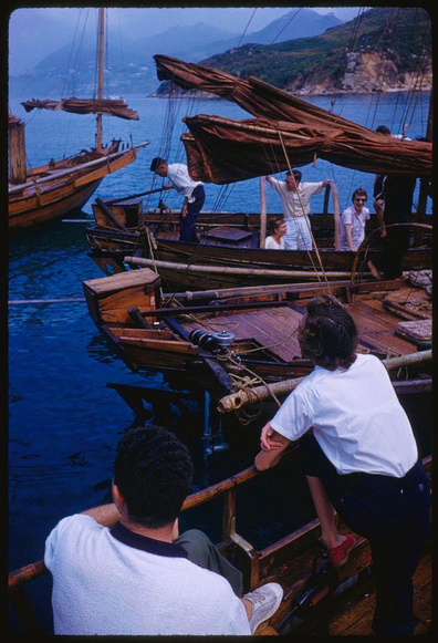 three wooden boats with people on them in a body of water. the boat on the right has two women and one man sitting at it, while the middle ship has three men standing up working to adjust sail. there are more ships off into distance but these ones appear closer as they take center stage within this photo.
