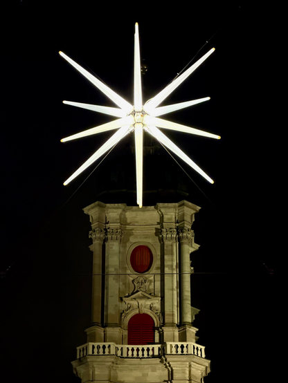 Eine hell erleuchtete Sternenskulptur sitzt nachts auf einem dekorativen Gebäude. Der Stern hat zahlreiche spitze Strahlen, die nach außen strahlen und sich vom dunklen Himmel abheben. Das Gebäude zeichnet sich durch einen klassischen Architekturstil mit roten Akzenten aus, was zum nächtlichen Ambiente beiträgt.