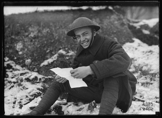 The image depicts an individual seated in a snowy landscape, likely during or after World War I. The person is wearing military attire typical of that era, including a helmet and overcoat, suggesting they are a soldier. They have glasses on their face and are smiling while holding what appears to be a piece of paper or letter. Their right hand seems slightly deformed at the wrist, possibly indicating some injury from combat.

The backdrop consists of snow-covered ground with patches of exposed earth and low vegetation, indicative of winter conditions in an outdoor setting like a battlefield or training grounds during that period. The overall tone is one of resilience and camaraderie amidst harsh circumstances.