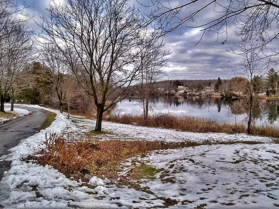 Snow covers the ground of a park next to a river. Orange and yellow grass can be seen sticking up through the snow and a curved paved pathway is seen on the left. Large leafless trees are seen as well as the calm water of the river reflecting the distant shoreline. Moody clouds fill the sky.