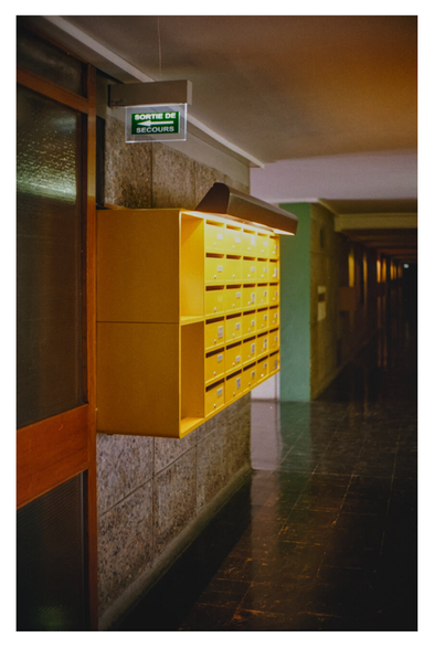 Color photograph of yellow mailboxes in the hallway of the Cité Radieuse, the apartment building designed by Le Corbusier in Marseille. The mailboxes appear to float and are illuminated from above by a beautiful light. Above the mailboxes, there is also a sign for the emergency exit (Sortie de secours), illuminated in green.