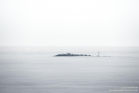 This image captures a serene and almost mystical scene of a mountain peak emerging from a sea of clouds. The landscape is shrouded in a thick layer of mist or fog, creating an ethereal and tranquil atmosphere. Only the top of the mountain and a few structures, possibly buildings or communication towers, are visible above the cloud cover.

The soft, muted colors and the smooth texture of the clouds give the scene a dreamy quality. The contrast between the solid structures and the fluid, cloudy background adds a sense of depth and mystery. This kind of view is often seen from high altitudes or during early mornings when clouds fill the valleys, creating an enchanting and peaceful ambiance. It evokes a sense of calm and wonder, highlighting the beauty and majesty of nature.