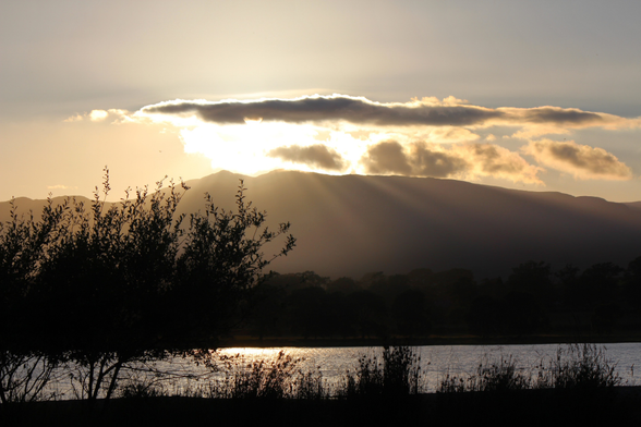 Beyond the lake in front of us is a mountain top, behind which the sun is setting. A flurry of clouds breaks the rays into beams that highlight the mountainside and make silhouettes of the trees below. The light reflects on the water's surface. Bala, Wales, UK. 