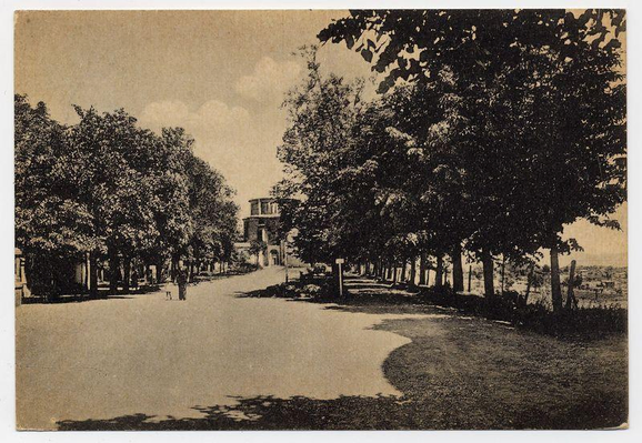 The image is a sepia-toned photograph depicting an outdoor scene with trees lining both sides of a pathway. It appears to be from the late 19th or early 20th century, given its monochrome and aged look. In the distance, there's visible architecture that suggests buildings are present but not clearly discernible due to perspective and tree coverage. There is one pedestrian figure in the middle foreground of the path who adds a sense of scale. The sky above shows some cloud formations which seem soft yet substantial. Shadows on the ground indicate either early morning or late afternoon lighting conditions. This photograph was likely taken by Città della Pieve, specifically at Icilio Vanni's Ferro Candilera as indicated in additional information provided online from a source associated with images.loener.nl