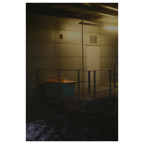 Versorgungsraum einer Schwebebahnhaltestelle, welche quer über einen Fluss (wupper) gebaut wurde. Eine einfache Metalltür führt von einer Plattform die über dem Wasser hängt in das graue Gebäude. Links von der Tür steht ein grüner Behälter mit Streusalz. Es ist dunkel. Die Szene wird von einer grünlich leuchtende Neonröhre oberhalb der Tür schwach ausgeleuchtet. 