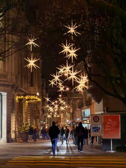 Eine geschäftige Stadtstraße bei Nacht, geschmückt mit hellen sternförmigen Lichtern. Die Menschen gehen den Bürgersteig entlang, und festliche Dekorationen sind auf den Gebäuden sichtbar, was eine fröhliche Atmosphäre schafft. Straßenschilder und Schaufenster sind beleuchtet und tragen zur pulsierenden Szene bei.