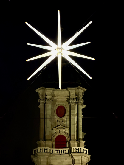 Eine hell erleuchtete Sternenskulptur sitzt nachts auf einem dekorativen Gebäude. Der Stern hat zahlreiche spitze Strahlen, die nach außen strahlen und sich vom dunklen Himmel abheben. Das Gebäude zeichnet sich durch einen klassischen Architekturstil mit roten Akzenten aus, was zum nächtlichen Ambiente beiträgt.