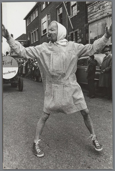 The image shows a person in the foreground with arms wide open, wearing an oversized striped garment that resembles both a sweater and skirt. The individual is also sporting high-top sneakers with laces tied into bows at the top of each shoe. Behind this central figure are other people gathered on what appears to be a street setting during daylight hours. In the background stands another person observing from behind. Several cars, including one labeled "Haaf" in Dutch, and text reading "usschemig," which is likely misspelled or misinterpreted German for 'schwergewicht' meaning heavy weight, are visible. The environment suggests a public gathering possibly related to an event such as the mentioned Carnaval celebration.