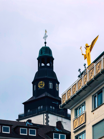 Blick nach oben in der Stadt Gießen: Dächer und Wohnhäuser rahmen den Turm der Stadtkirche, daneben ein Gebäude mit goldener Figur auf dem Dach, vor hellem, ruhigem Himmel.