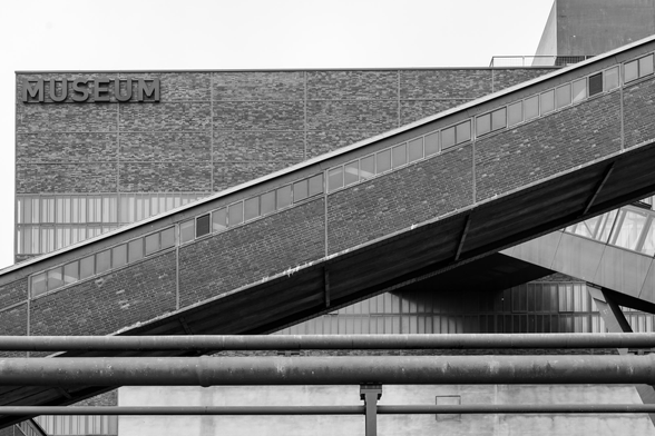 Black-and-white photo of the Zollverein Coal Mine Industrial Complex. In the foreground, former conveyor belts run diagonally across the image, while the rectangular museum building can be seen in the background. All of the buildings are made of brick.