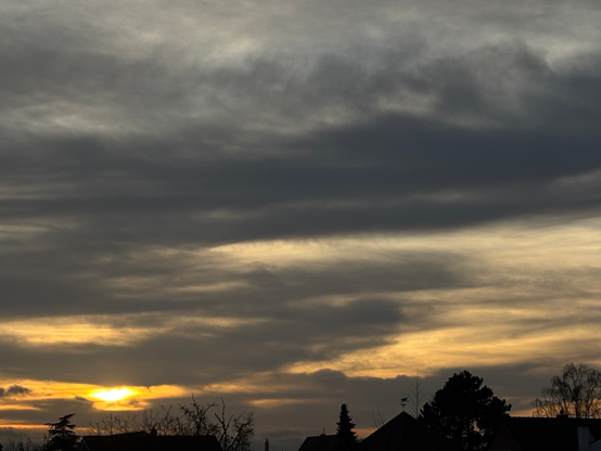 Am unteren rechten Bildrand zeigt sich die Silhouette eines größeren Baumes sowie von Sträuchern und Dächern. Von unten links verteilt sich das letzte Licht der untergehenden Sonne, die selbst links als kleiner verschwommener goldener Ball leuchtet. Das Licht sendet golden Farben über den gesamten Himmels. Der Himmel ist teilweise langgezogen grau bewölkt.