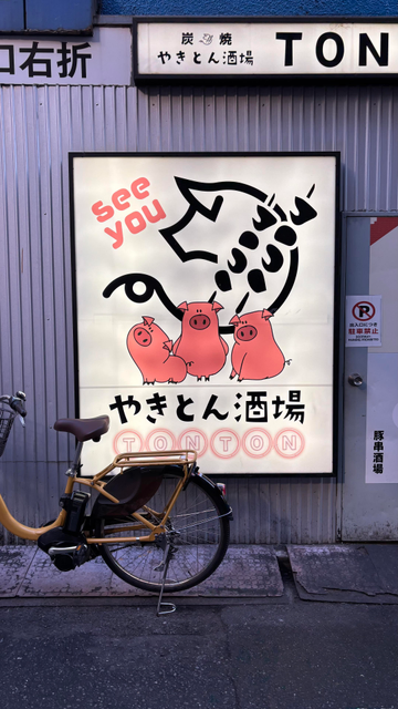 side of a restaurant. the wall is covered in corrugated metal with vertical lines. on the wall, a large poster-sized illuminated illustration "see you" with three piglets and the restaurant name TONTON. in the top right part of an illuminated horizontal light with just TON visible. bottom right half of a parked bicycle