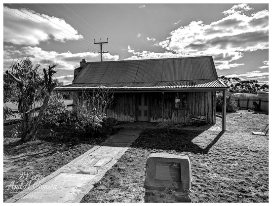 A black and white photograph of the historic Orroroo Early Settlers Cottage, a weathered wooden structure with a corrugated iron roof and a verandah, set beneath a dramatic sky with power lines visible overhead. A stone marker is in the foreground.